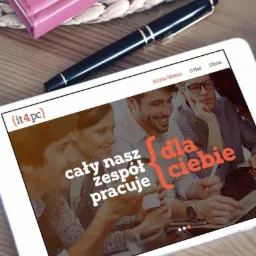 Tablet wyświetla stronę internetową firmy it4pc z hasłem 'Cały nasz zespół pracuje dla Ciebie', obok leży notatnik i długopis na drewnianym biurku.