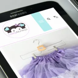 Tablet wyświetla stronę internetową z logo LovelyMonkeys i zdjęciem fioletowej tiulowej spódniczki dziecięcej na wieszaku.