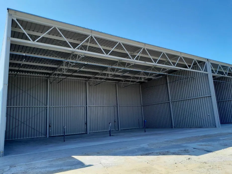 Nowoczesna hala z blachy trapezowej w jasnych odcieniach szarości, z widoczną konstrukcją stalową dachu i betonową posadzką, w słoneczny dzień.