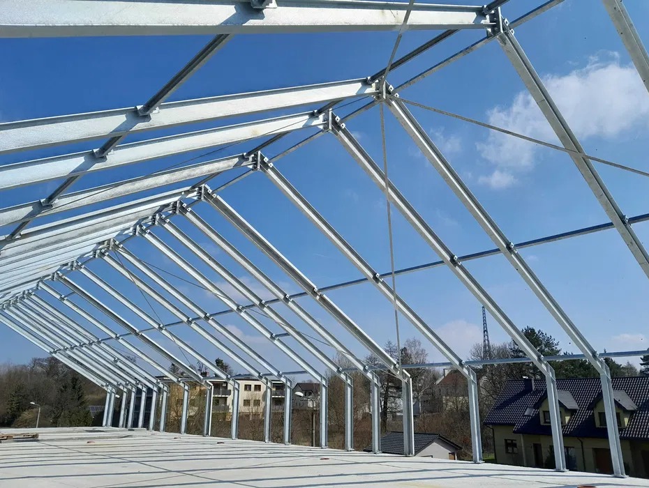 Szara, metalowa konstrukcja hali w trakcie budowy, widok z dołu na tle błękitnego nieba. Elementy złączone śrubami. W tle zabudowa jednorodzinna i drzewa.