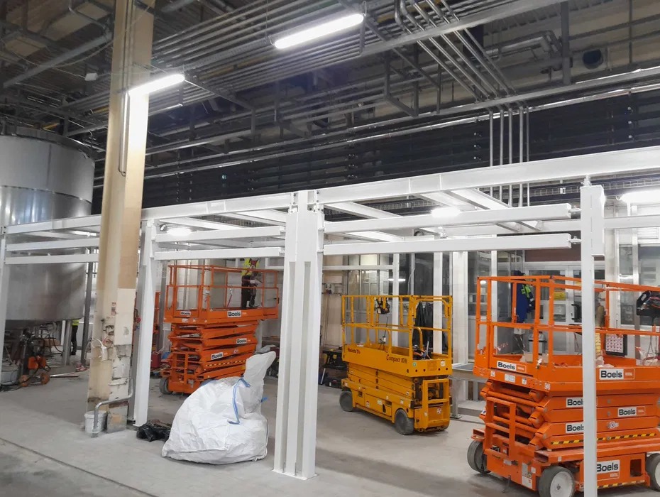 Montaż białej konstrukcji stalowej w hali przemysłowej. Widoczne podnośniki koszowe i pracownicy w odblaskowych ubraniach. Rury pod sufitem.
