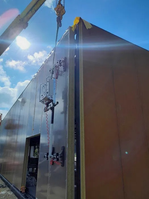 Montaż paneli warstwowych na hali z użyciem przyssawki próżniowej podwieszonej na dźwigu, widoczne łączenie paneli i niebo z chmurami w tle.