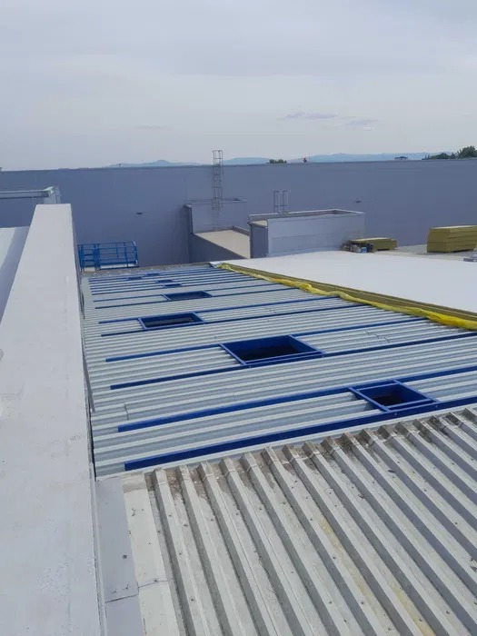 Szary dach hali z blachy trapezowej z niebieskimi obramowaniami otworów okiennych, widok z góry, w tle biała ściana hali i drabina.