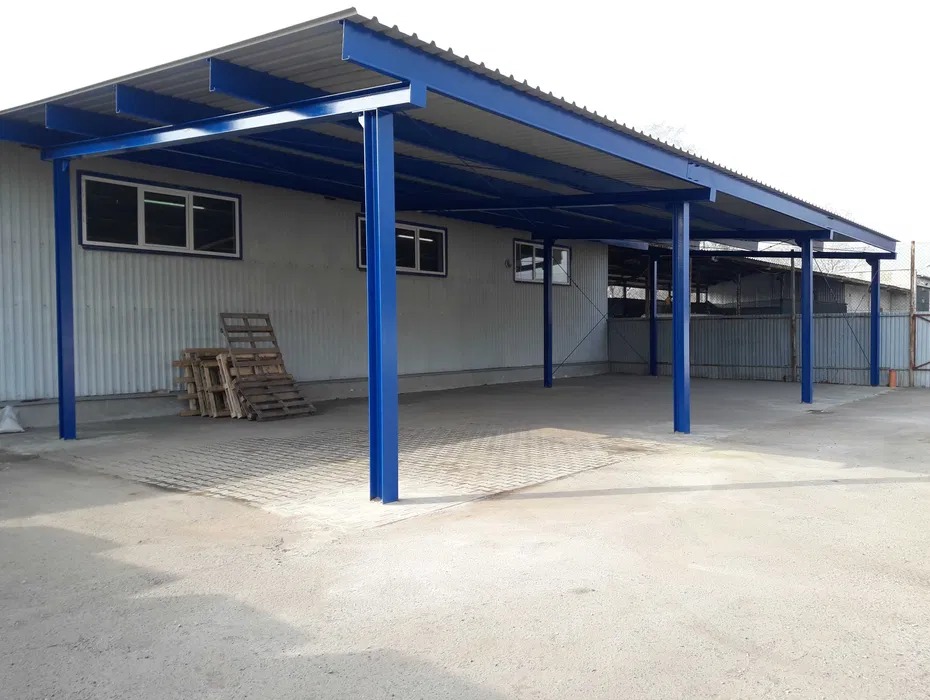 Zadaszenie z niebieską konstrukcją stalową przy budynku z blachy trapezowej. Widoczne palety i kostka brukowa na posadzce.