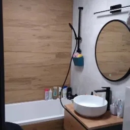 Nowoczesna łazienka z wanną obłożoną panelami drewnopodobnymi, okrągłym lustrem z podświetleniem LED, umywalką nablatową i czarną armaturą.