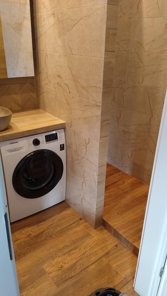 Ukończona łazienka z pralką Samsung, drewnopodobną podłogą i płytkami imitującymi marmur na ścianach, widoczny fragment kabiny prysznicowej z niskim progiem.