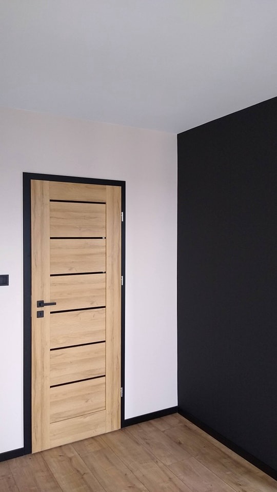 Wnętrze po remoncie: Jasne drewnopodobne drzwi z czarnymi akcentami na tle białej i grafitowej ściany, wykończone czarnymi listwami przypodłogowymi i panelami podłogowymi.