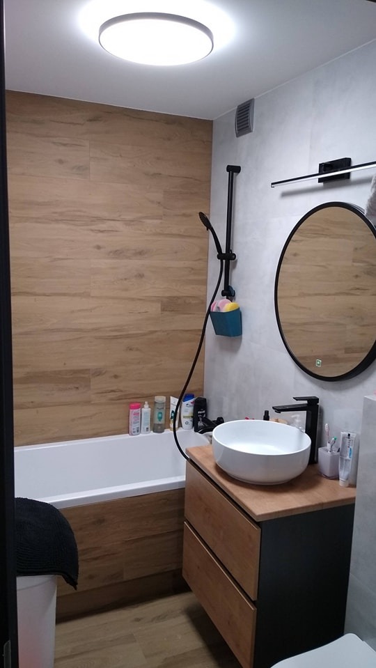 Nowoczesna łazienka z wanną obłożoną panelami drewnopodobnymi, okrągłym lustrem z podświetleniem LED, umywalką nablatową i czarną armaturą.