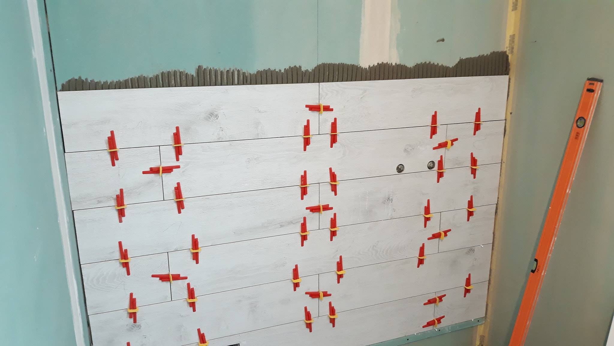 Ściana w trakcie układania płytek imitujących drewno, z widocznymi czerwonymi krzyżykami dystansowymi i poziomicą po prawej stronie.