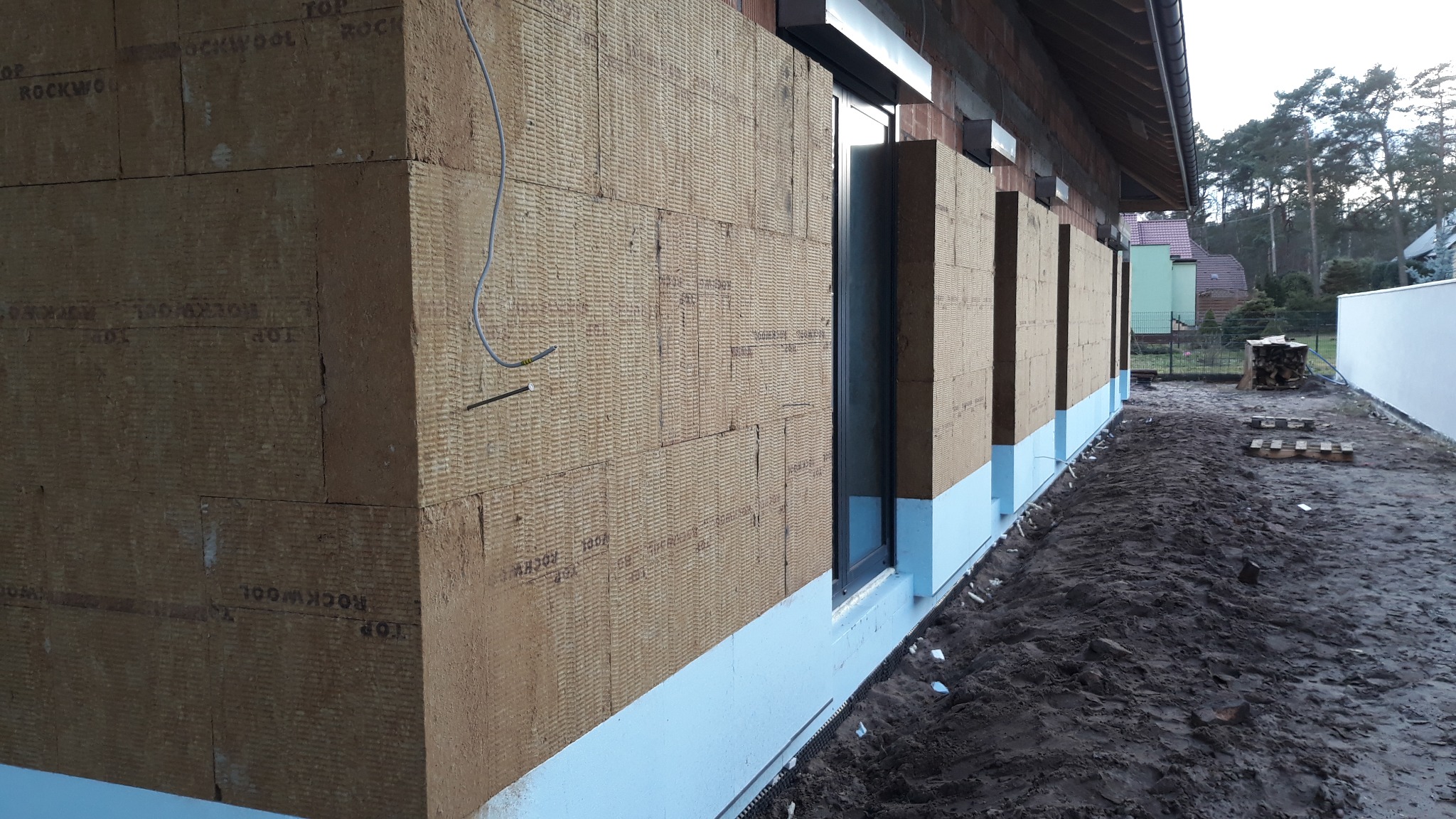 Ocieplenie zewnętrzne budynku z widoczną warstwą wełny mineralnej Rockwool i niebieską izolacją fundamentów, z fragmentem okna i instalacją elektryczną.
