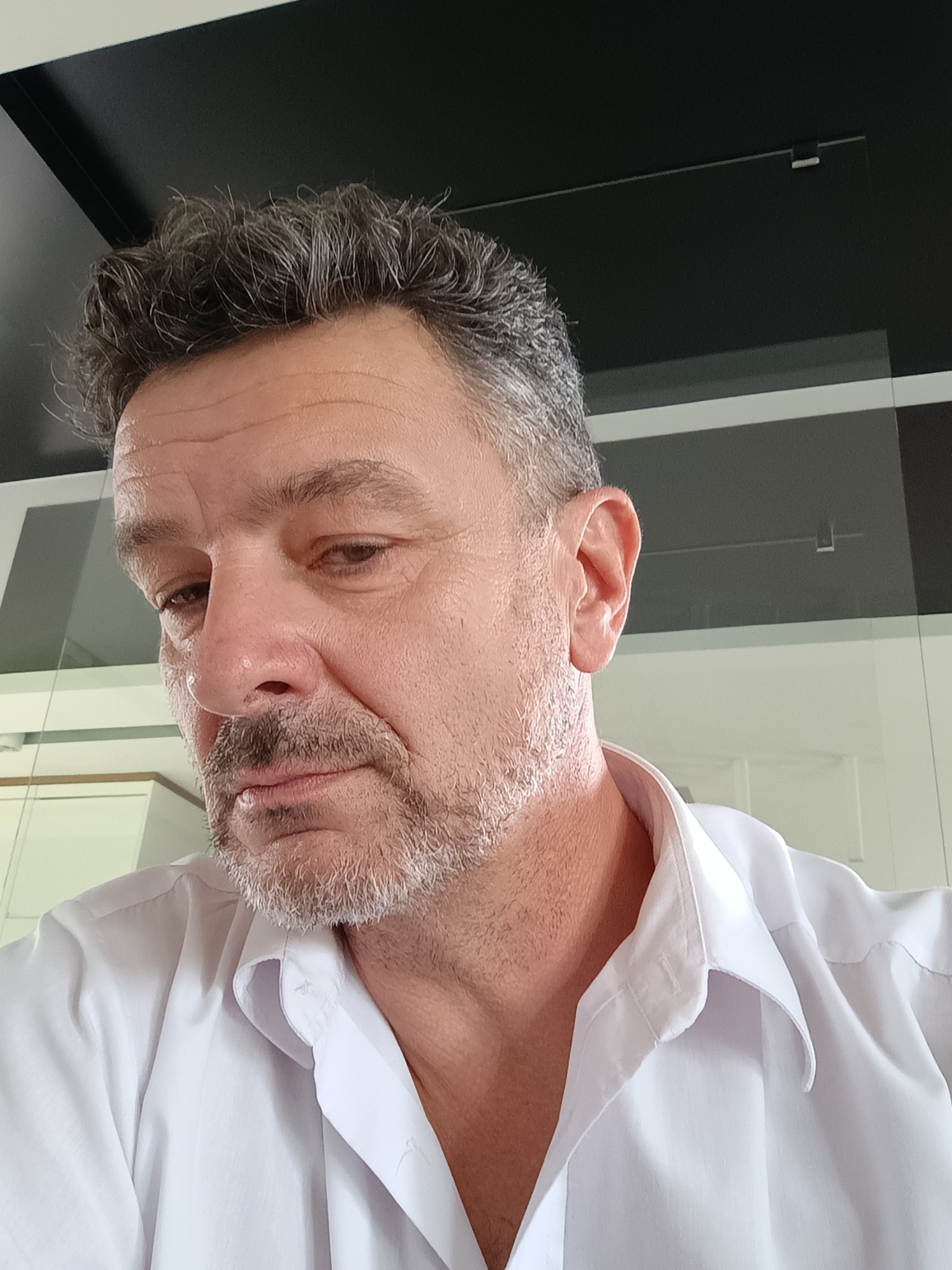 Portret mężczyzny w białej koszuli, z krótką brodą i zmarszczkami mimicznymi, uchwycony z góry, z widocznym fragmentem nowoczesnego wnętrza w tle.