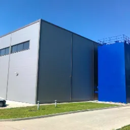 Nowoczesny budynek przemysłowy z szarej blachy trapezowej z niebieskim zbiornikiem na zewnątrz, widziany w słoneczny dzień z samochodem w tle.