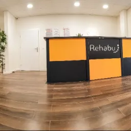 Jasne wnętrze poczekalni gabinetu rehabilitacyjnego z pomarańczową sofą, zegarem ściennym, roślinami doniczkowymi i recepcją z logo 'Rehabu' w kolorach pomarańczowo-czarnych, widok na zewnątrz...