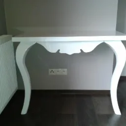 Białe biurko w stylu ludwikowskim z błyszczącym blatem i giętymi nogami, na tle szarej ściany z gniazdkami elektrycznymi i ciemnej podłogi.