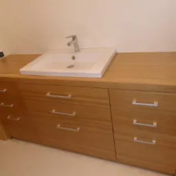 Łazienkowa szafka pod umywalkę z drewnianym blatem i trzema szufladami z poziomymi, metalowymi uchwytami, biała umywalka ceramiczna z chromowaną baterią.