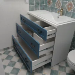 Biała szafka łazienkowa z umywalką i trzema wysuwanymi, błękitnymi szufladami z ozdobnymi uchwytami, na tle wzorzystej, zielono-białej glazury i toalety.