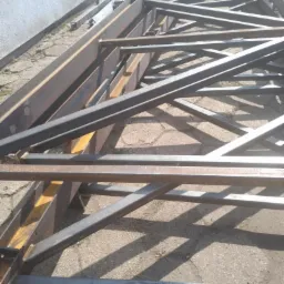 Konstrukcja stalowa z profili, spawana, w surowym stanie, leżąca na ziemi. Widoczne spawy i niejednolita kolorystyka stali. Element budowy hali.