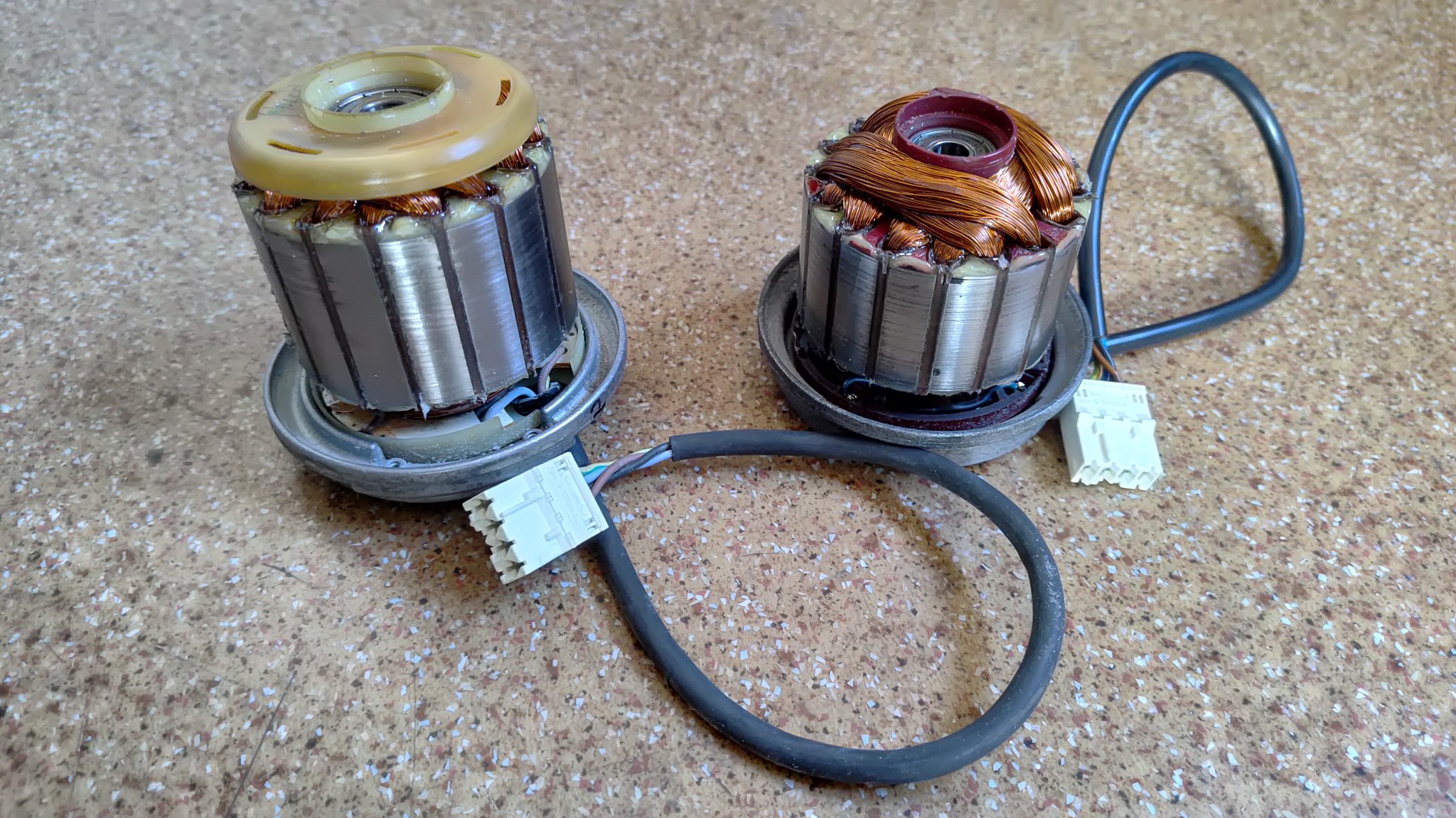Dwa wirniki elektronarzędzi z uzwojeniami miedzianymi i plastikowymi elementami, leżące na wzorzystej powierzchni. Widoczne przewody i złącza.