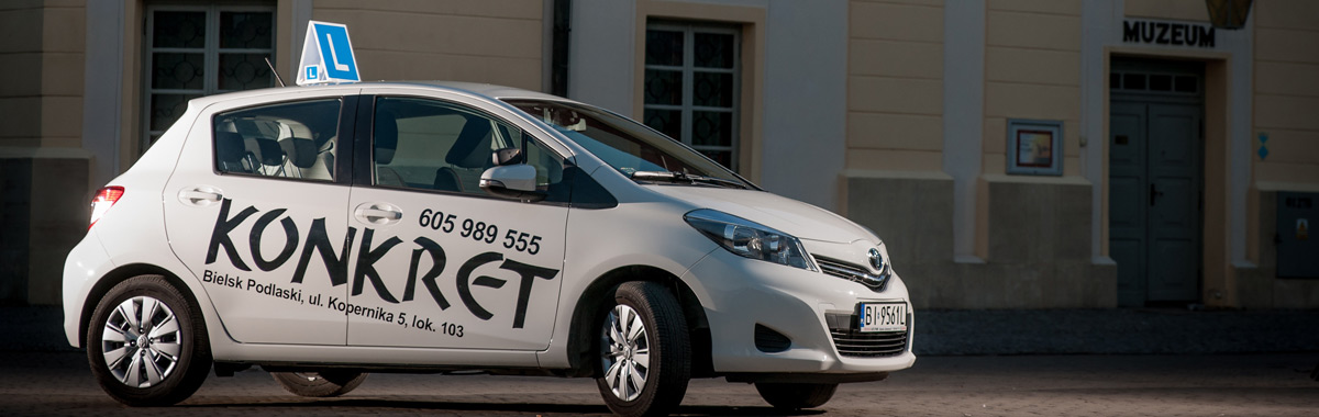 Biały samochód z oznaczeniem 'L' na dachu i napisem 'KONKRET' oraz numerem telefonu na boku, zaparkowany przed budynkiem z napisem 'MUZEUM'.