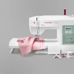 Nowoczesna, biała maszyna do szycia Singer Brilliance z różową tkaniną w białe kropki pod igłą, obok leżą nożyczki, szpulki nici i miarka krawiecka.