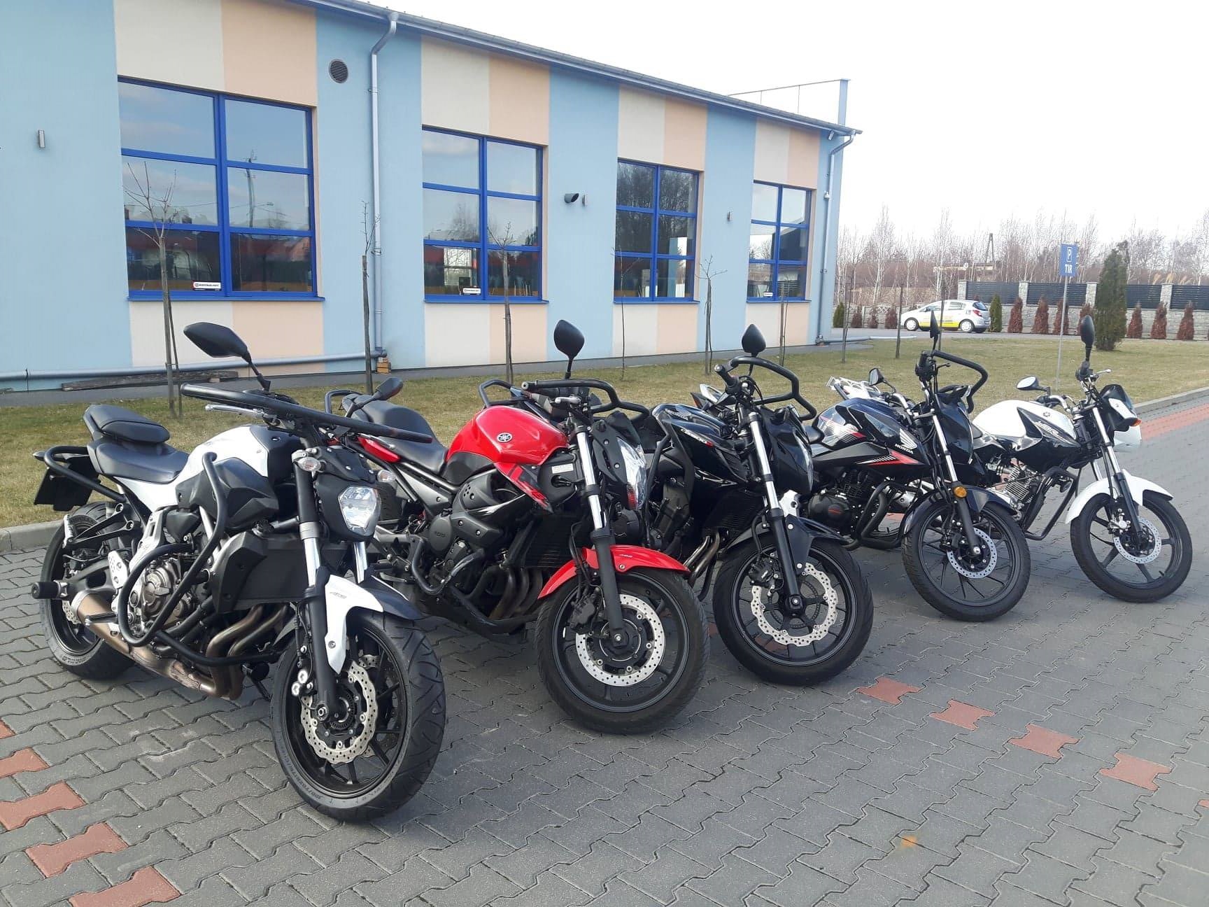 Rząd motocykli różnych marek i kolorów zaparkowanych przed budynkiem w Mińsku Mazowieckim; widoczne motocykle białe, czerwone i czarne.