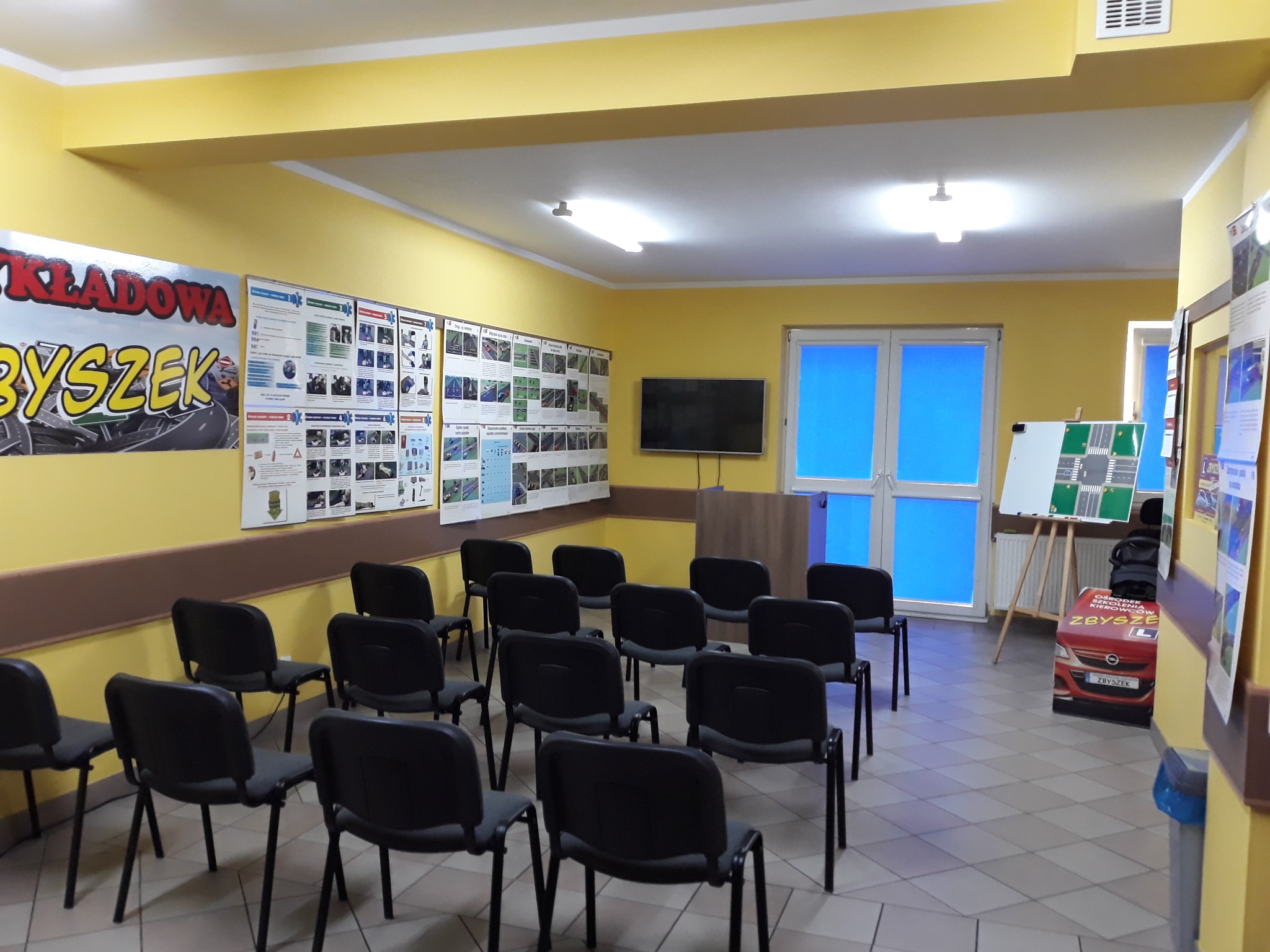 Sala wykładowa z krzesłami, plakatami tematycznymi na żółtych ścianach, tablicą i kartonowym modelem samochodu z napisem 'Zbyszek'.