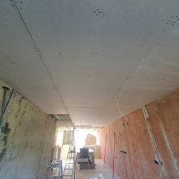 FACH- LUX Jeremiasz Kolankiewicz - Sufit podwieszany z płyt kartonowo-gipsowych w trakcie montażu, widoczne łączenia płyt i otwory na oświetlenie, ściany przygotowane pod wykończenie, drabina i narzędzia w tle.