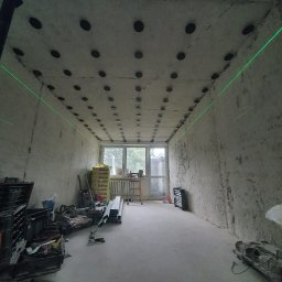 FACH- LUX Jeremiasz Kolankiewicz - Przygotowanie do montażu sufitu podwieszanego: surowy betonowy strop z nawierconymi otworami i zamocowanymi kołkami, wyznaczony poziom zielonym laserem, w tle okno i materiały izolacyjne,...