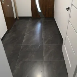 Ułożona, ciemnoszara podłoga z płytek imitujących kamień w przedpokoju, widoczne drzwi wejściowe z elementami drewnopodobnymi i szklanymi, oraz białe drzwi wewnętrzne po prawej stronie.