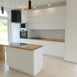 Jasna kuchnia z białymi szafkami, drewnianym blatem i wyspą kuchenną z płytą indukcyjną, widok z okna na zieleń.