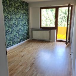 FACH- LUX Jeremiasz Kolankiewicz - Pusty pokój po remoncie z drewnianą podłogą, ścianą w ciemnozieloną tapetę z motywem roślinnym oraz otwartym oknem balkonowym z widokiem na zieleń.