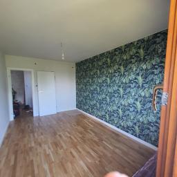 FACH- LUX Jeremiasz Kolankiewicz - Pokój po remoncie: parkiet ułożony, jedna ściana z tapetą w motywy roślinne, widoczne drzwi wejściowe i drzwi do kolejnego pomieszczenia.