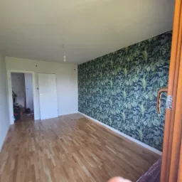 Pokój po remoncie: parkiet ułożony, jedna ściana z tapetą w motywy roślinne, widoczne drzwi wejściowe i drzwi do kolejnego pomieszczenia.