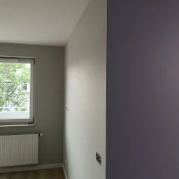 Świeżo pomalowane wnętrze z dwiema odmiennymi barwami ścian: szarą przy oknie i ciemnofioletową na sąsiedniej ścianie, widoczny grzejnik, surowe gniazdka elektryczne i fragment podłogi z paneli.