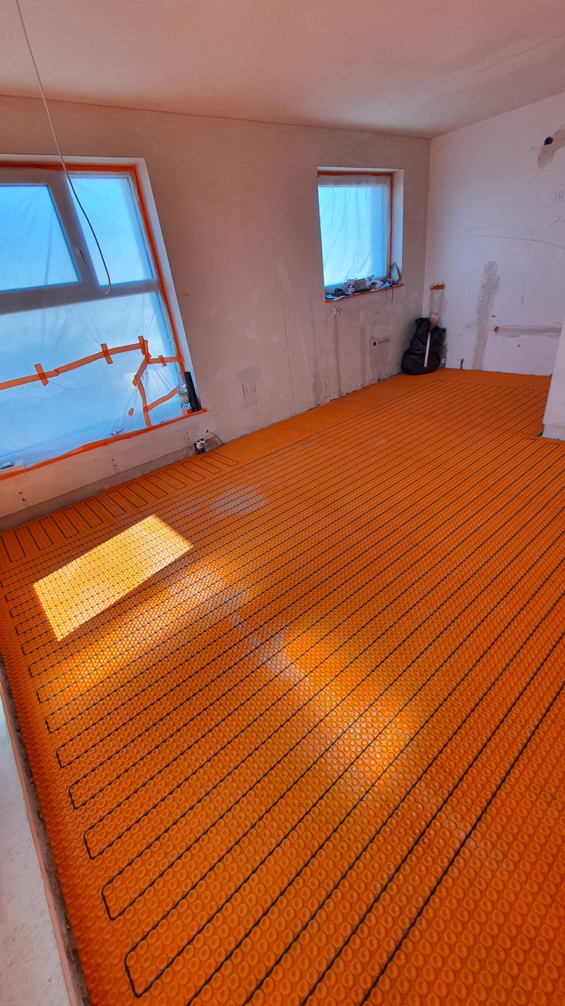 Pomarańczowa mata grzewcza podłogowa, ułożona w surowym wnętrzu z oknami zabezpieczonymi folią, przygotowana do wylewki.