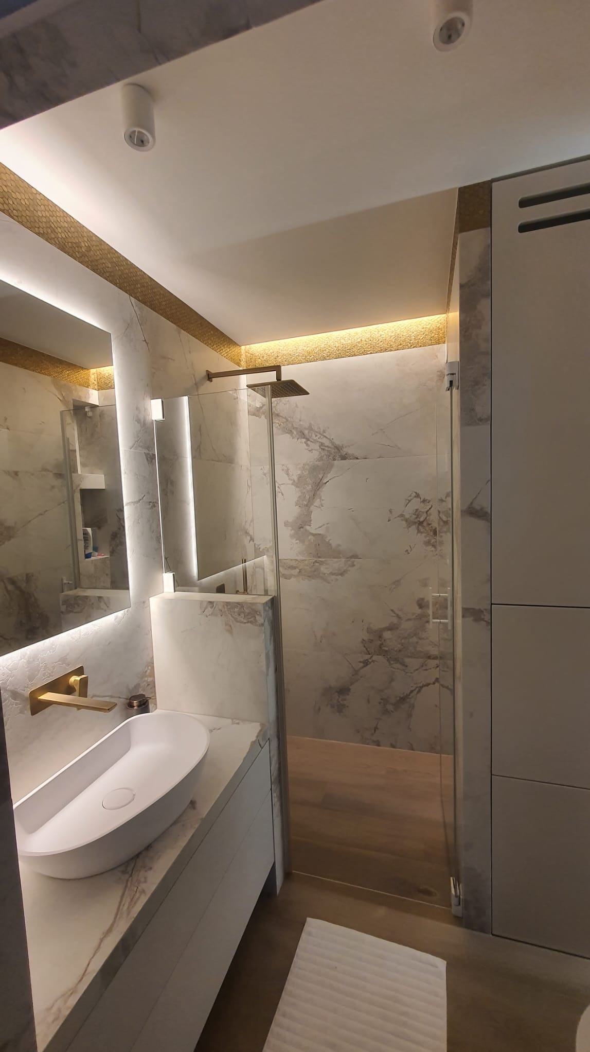 Nowoczesna łazienka w Świebodzinie z marmurowymi płytkami, złotą armaturą i podświetlanym lustrem, ukazująca kabinę prysznicową typu walk-in i umywalkę nablatową.