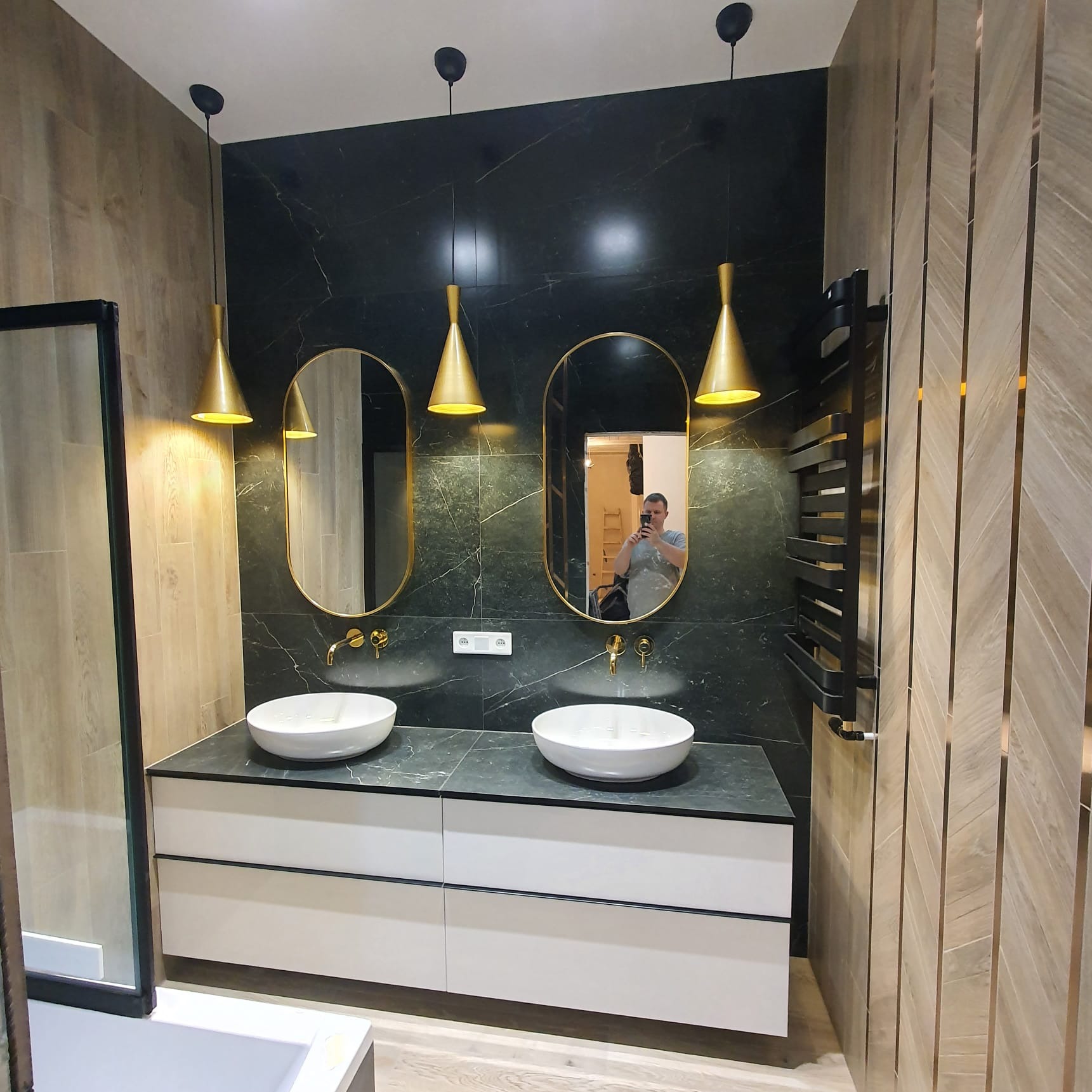 Nowoczesna łazienka z czarną marmurową ścianą, dwoma owalnymi lustrami w złotych ramach, białymi umywalkami nablatowymi, złotą armaturą, białymi szafkami pod umywalkami i złotymi lampami wiszącymi...
