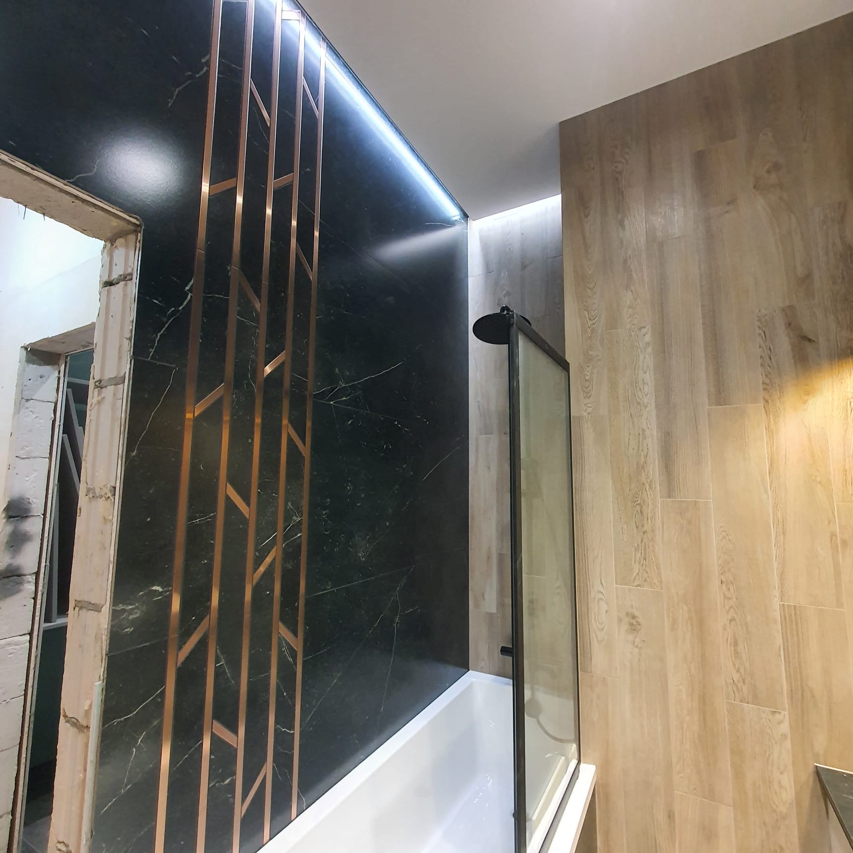Wykończona łazienka z czarną ścianą z marmuru z miedzianymi dekoracyjnymi listwami, kabiną prysznicową typu walk-in i drewnopodobnymi płytkami na ścianie, widoczny surowy fragment ściany przed...