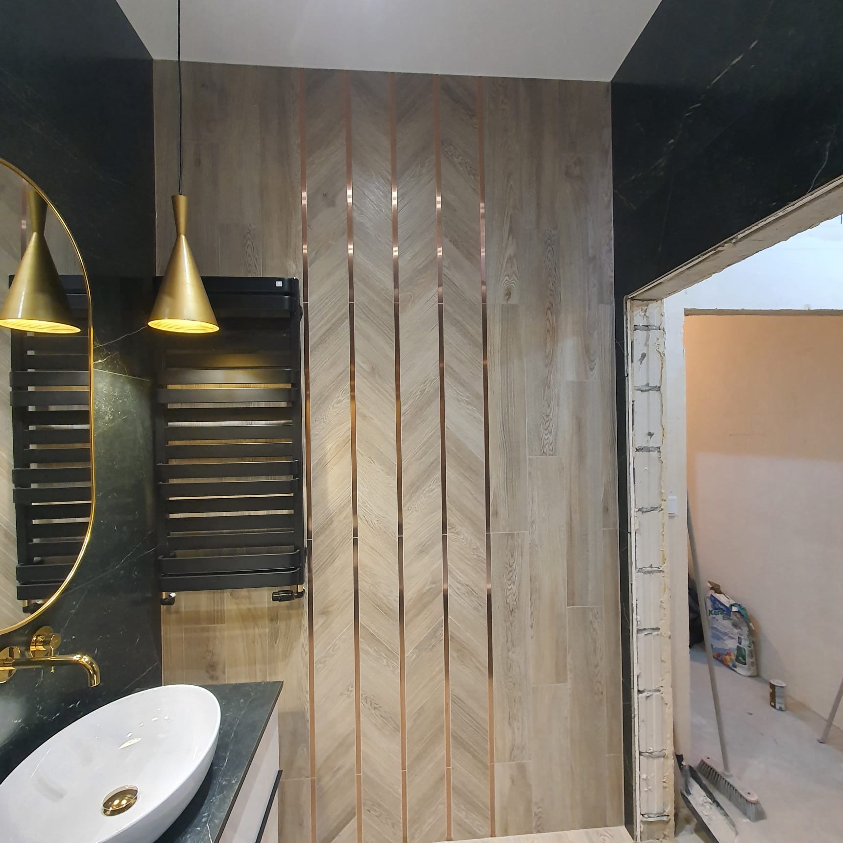 Nowoczesna łazienka w trakcie wykańczania: ściana z płytek imitujących drewno z pionowymi wstawkami w kolorze miedzi, czarny grzejnik drabinkowy, umywalka nablatowa z czarnym blatem i złotą...