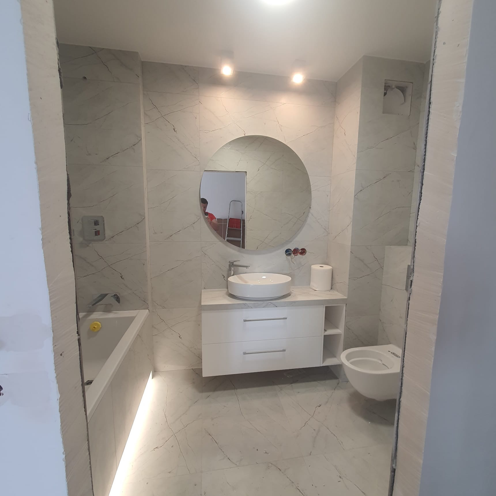 Wykończona łazienka z białymi płytkami imitującymi marmur, okrągłe lustro z odbiciem drabiny, biała szafka pod umywalkę z umywalką nablatową, podświetlana wanna, wisząca toaleta.