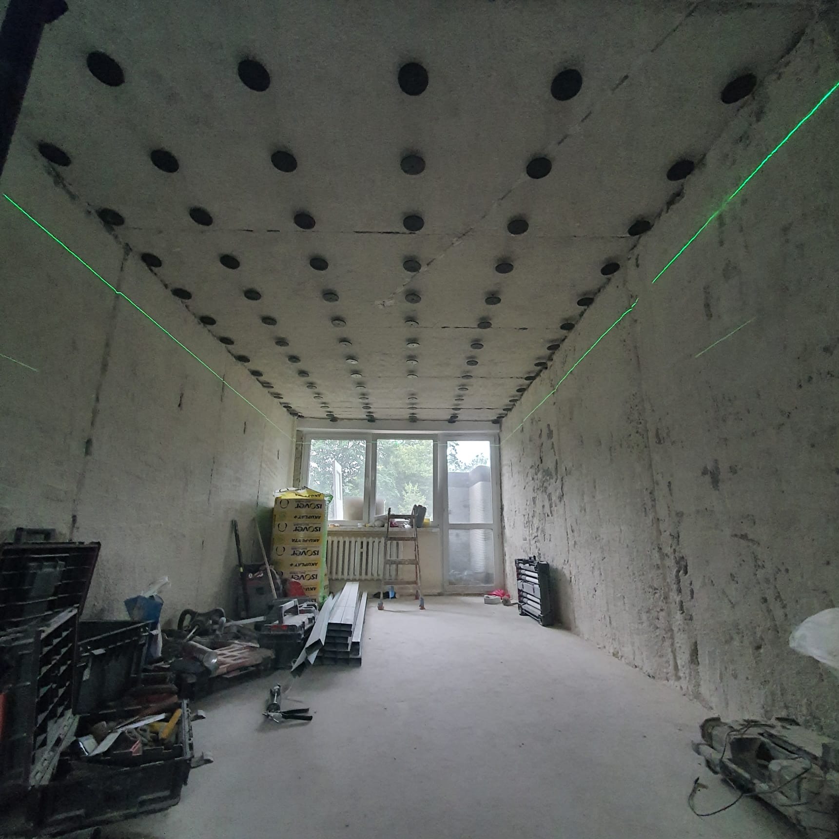 Przygotowanie do montażu sufitu podwieszanego: surowy betonowy strop z nawierconymi otworami i zamocowanymi kołkami, wyznaczony poziom zielonym laserem, w tle okno i materiały izolacyjne,...