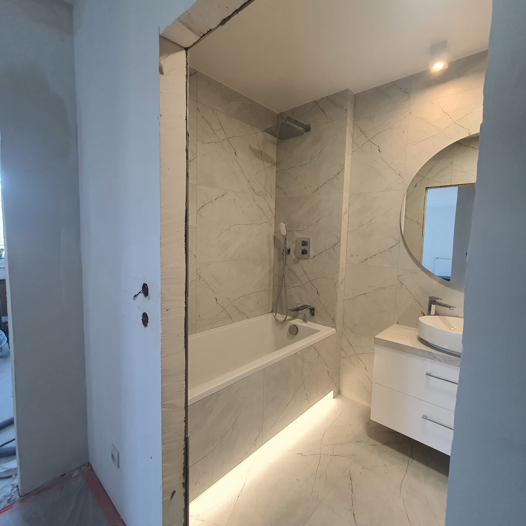 Nowoczesna łazienka w trakcie remontu: białe marmurowe płytki, wanna z podświetleniem LED, okrągłe lustro nad umywalką na szafce, surowe ściany w tle.