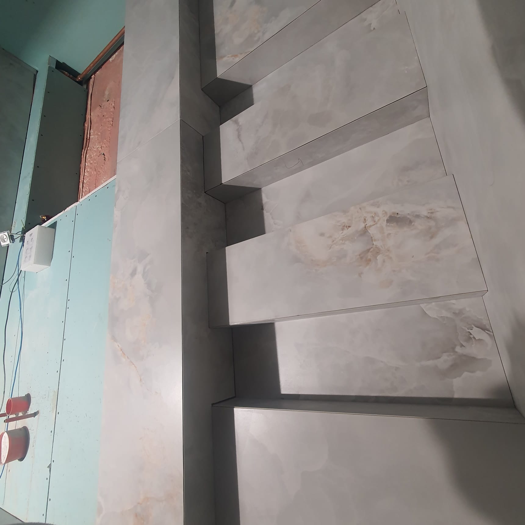 Wykończenie ściany z półkami z płytek imitujących marmur w trakcie prac, widoczna płyta kartonowo-gipsowa i elementy instalacji elektrycznej.