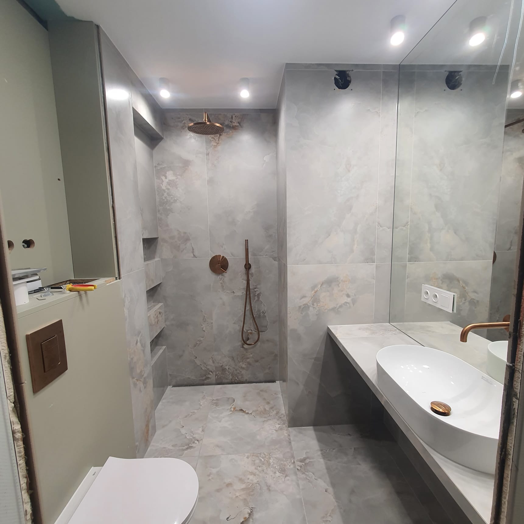 Nowoczesna łazienka z płytkami imitującymi marmur, armaturą w kolorze miedzi i umywalką nablatową o owalnym kształcie, widoczne niedokończone prace wykończeniowe przy zabudowie toalety.