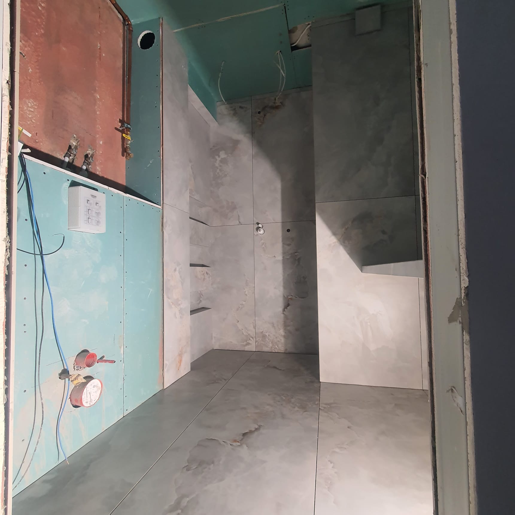 Wnętrze łazienki w trakcie remontu: ściany z płyt karton-gips, częściowo wykończone marmurowe płytki, widoczne instalacje wodne i elektryczne, surowy stan wykończenia.