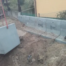 Montaż prefabrykowanych betonowych elementów fundamentowych z użyciem dźwigu, widoczne wykopane fundamenty i istniejące fragmenty muru oporowego z prefabrykatów.