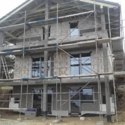 Dwukondygnacyjny dom w stanie surowym otwartym, z oknami i rusztowaniami, widoczne elementy konstrukcji dachu i bloczki betonu komórkowego.