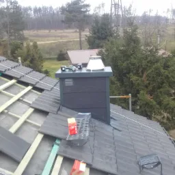 Montaż szarej dachówki na drewnianej konstrukcji dachu, widoczny komin z narzędziami oraz elementy wentylacyjne, w tle las i zabudowania.