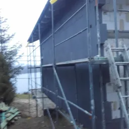 Budowa domu: elewacja z szarego styropianu na rusztowaniu, widoczna drabina i ściana z białych bloczków, w tle drzewa i jezioro.