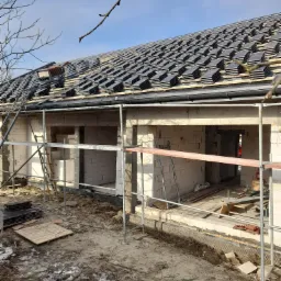 Wznoszony dom z białego bloczka, częściowo pokryty ciemną dachówką, widoczne rusztowania i elementy konstrukcyjne dachu, w tle błękitne niebo i gałęzie drzew